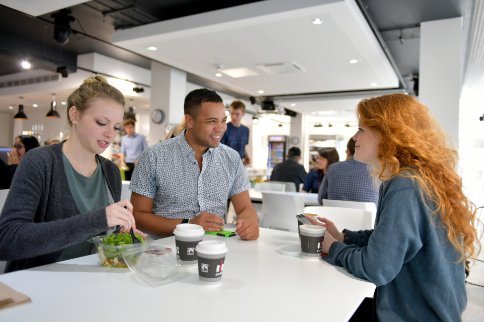 A female student, with ginger hair, smiling and chatting with a female student and a male student, having lunch.